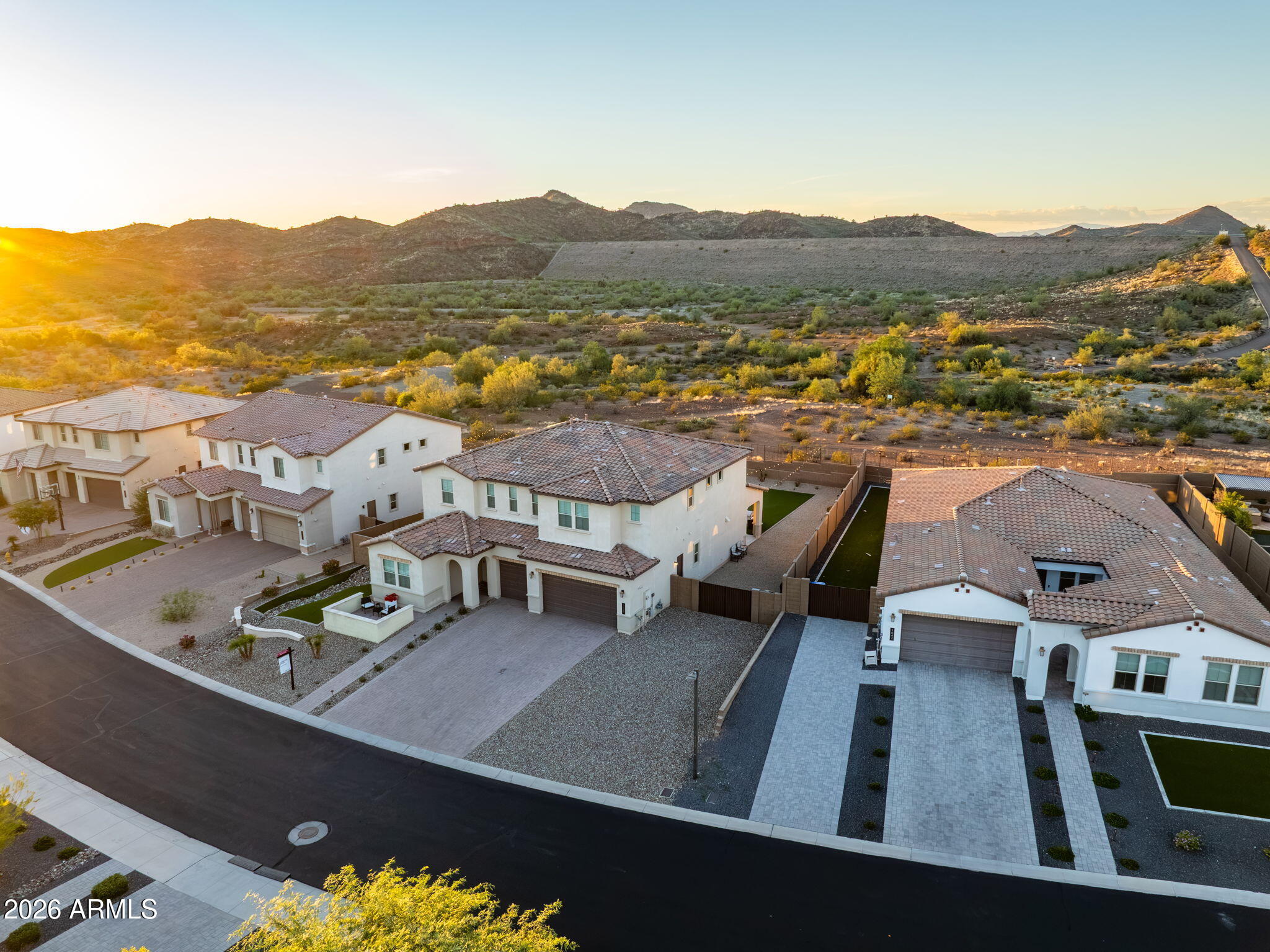 Aerial view of a stucco home with a tiled roof, neighboring houses, and desert mountains at sunset.