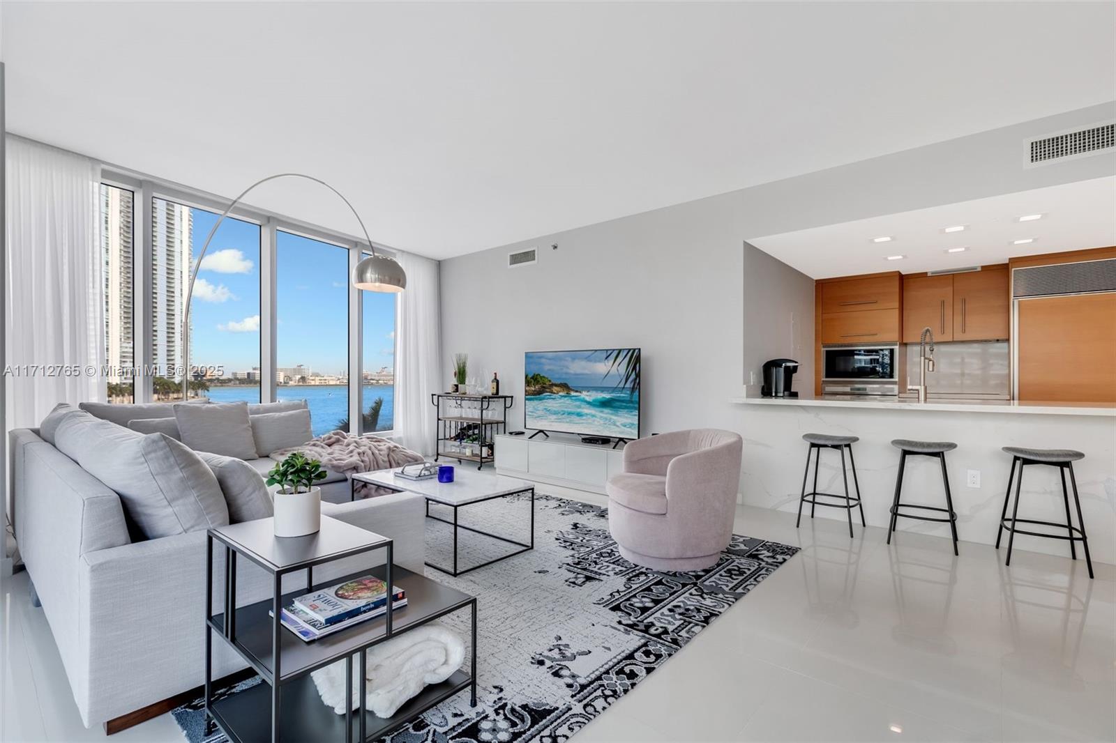 Bright furnished living room with floor-to-ceiling windows overlooking the water and an open kitchen.