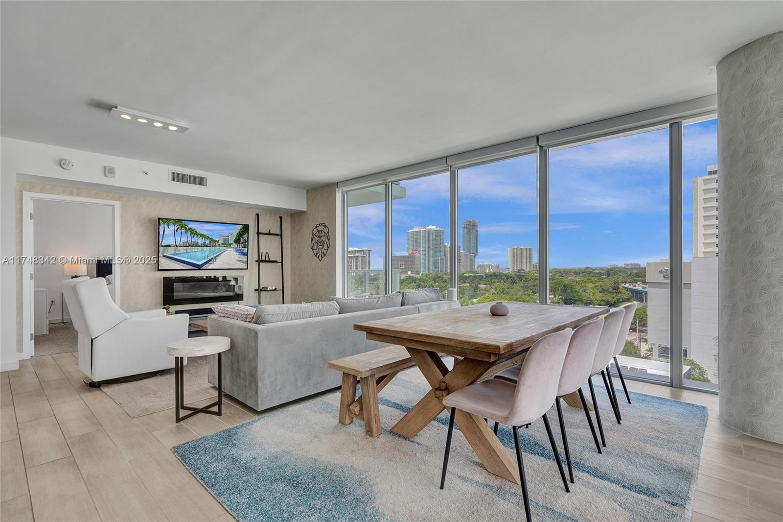 Living and dining room with floor-to-ceiling windows overlooking greenery and a city skyline.