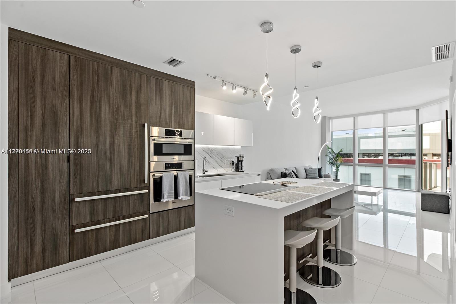 Modern kitchen with a large white island, dark wood cabinets, stainless appliances, and floor-to-ceiling windows.