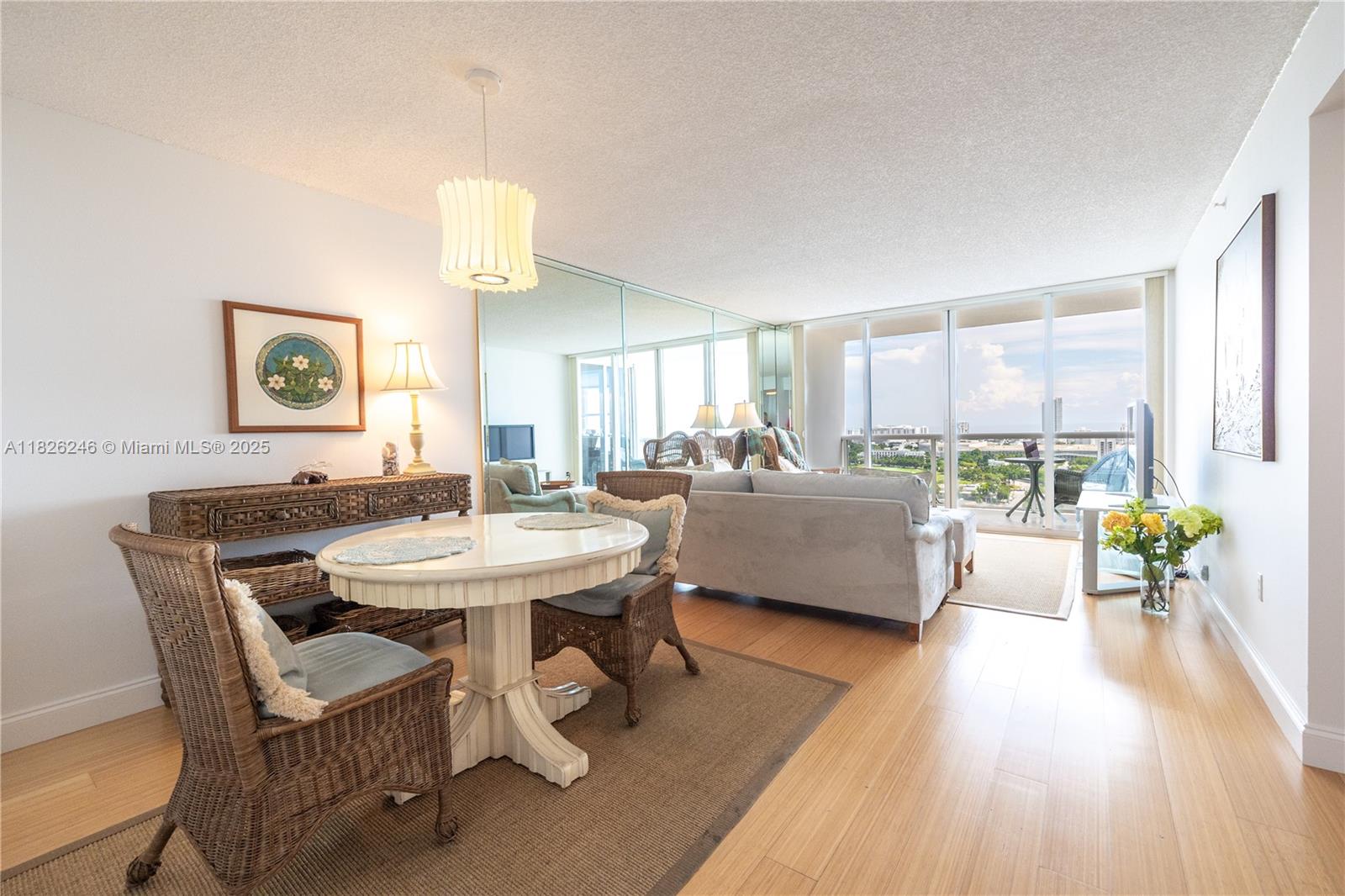 Furnished living and dining area with wicker chairs, mirrored divider, and balcony windows overlooking water.