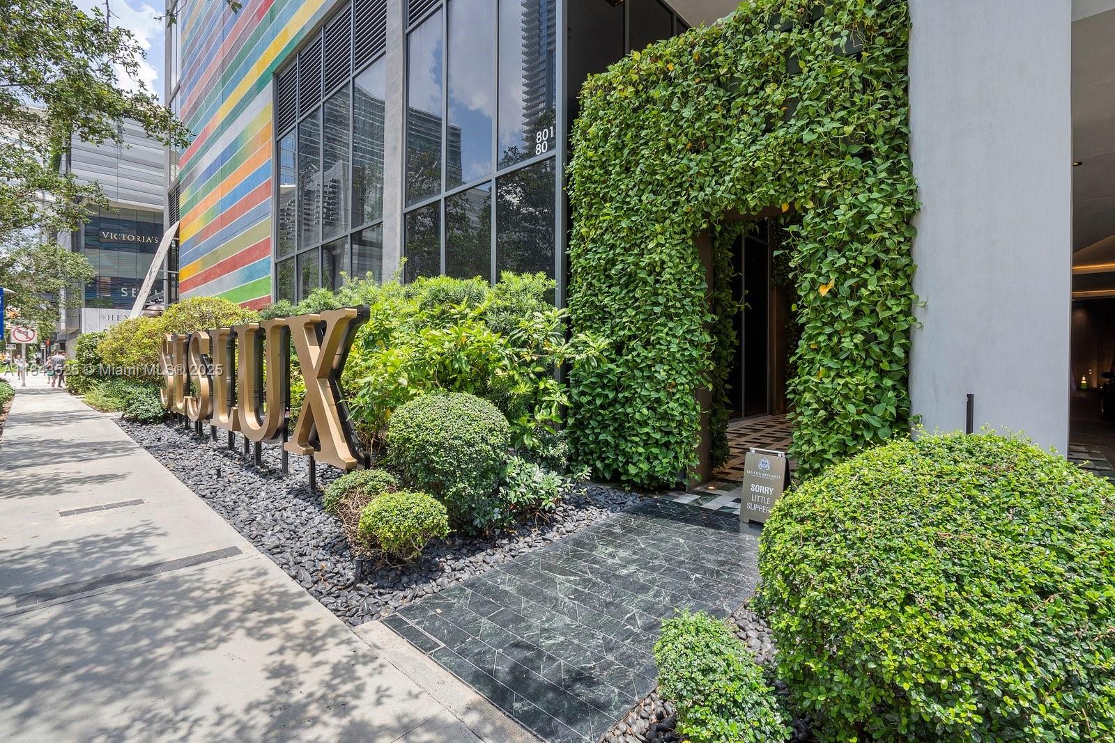 Modern building entrance framed by a leafy green wall, colorful facade bands, and landscaped shrubs.