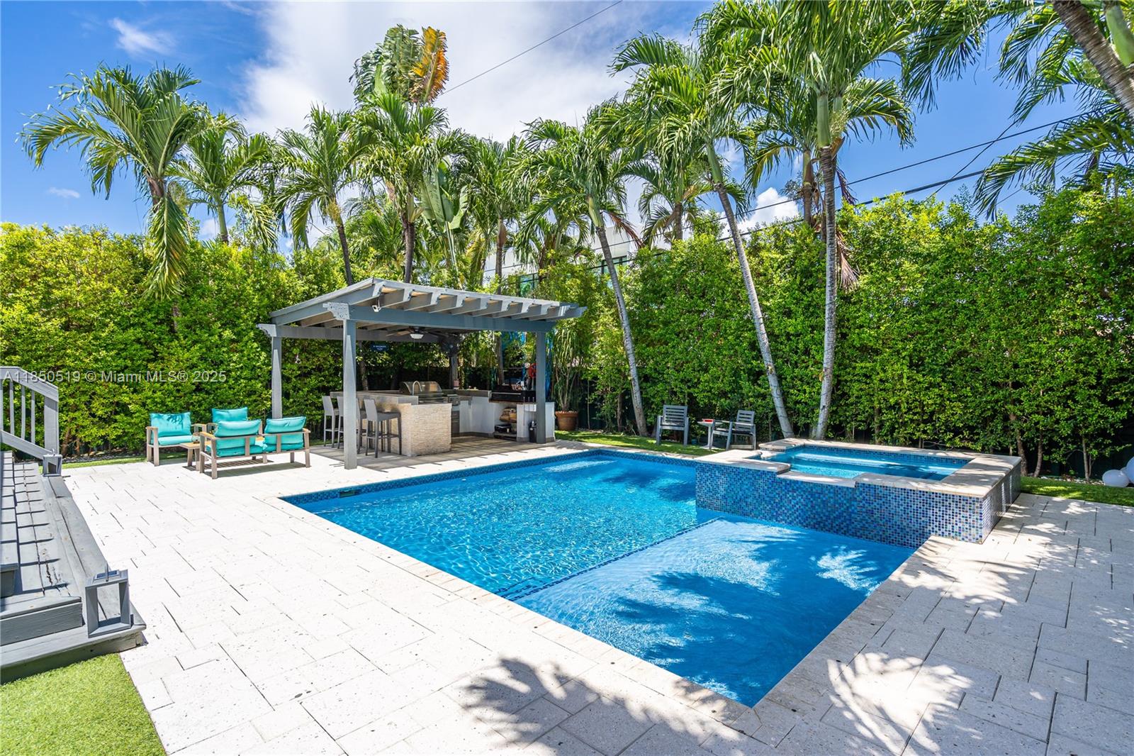 Backyard pool and raised spa beside a pergola-covered outdoor kitchen, palm trees, and turquoise lounge chairs.