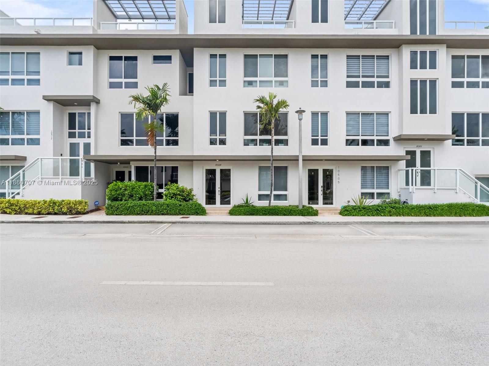 Modern white residential building with glass balconies, palm trees, and landscaped hedges.