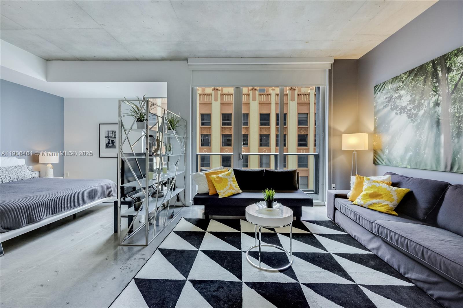 Modern furnished studio with a geometric rug, yellow pillows, and a neighboring building view.