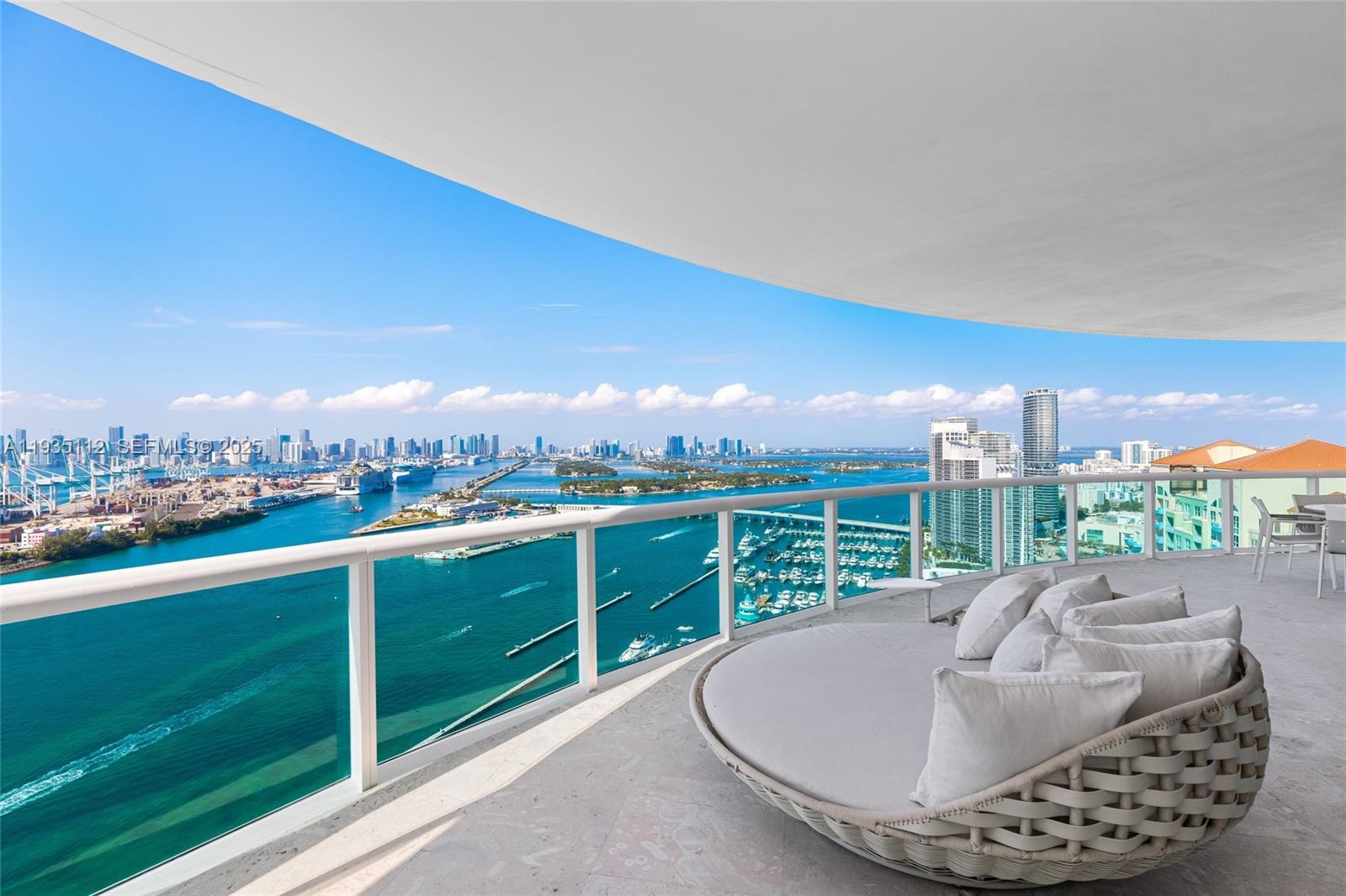 High-rise balcony with woven lounge seating overlooking turquoise water, a marina, and the Miami skyline.