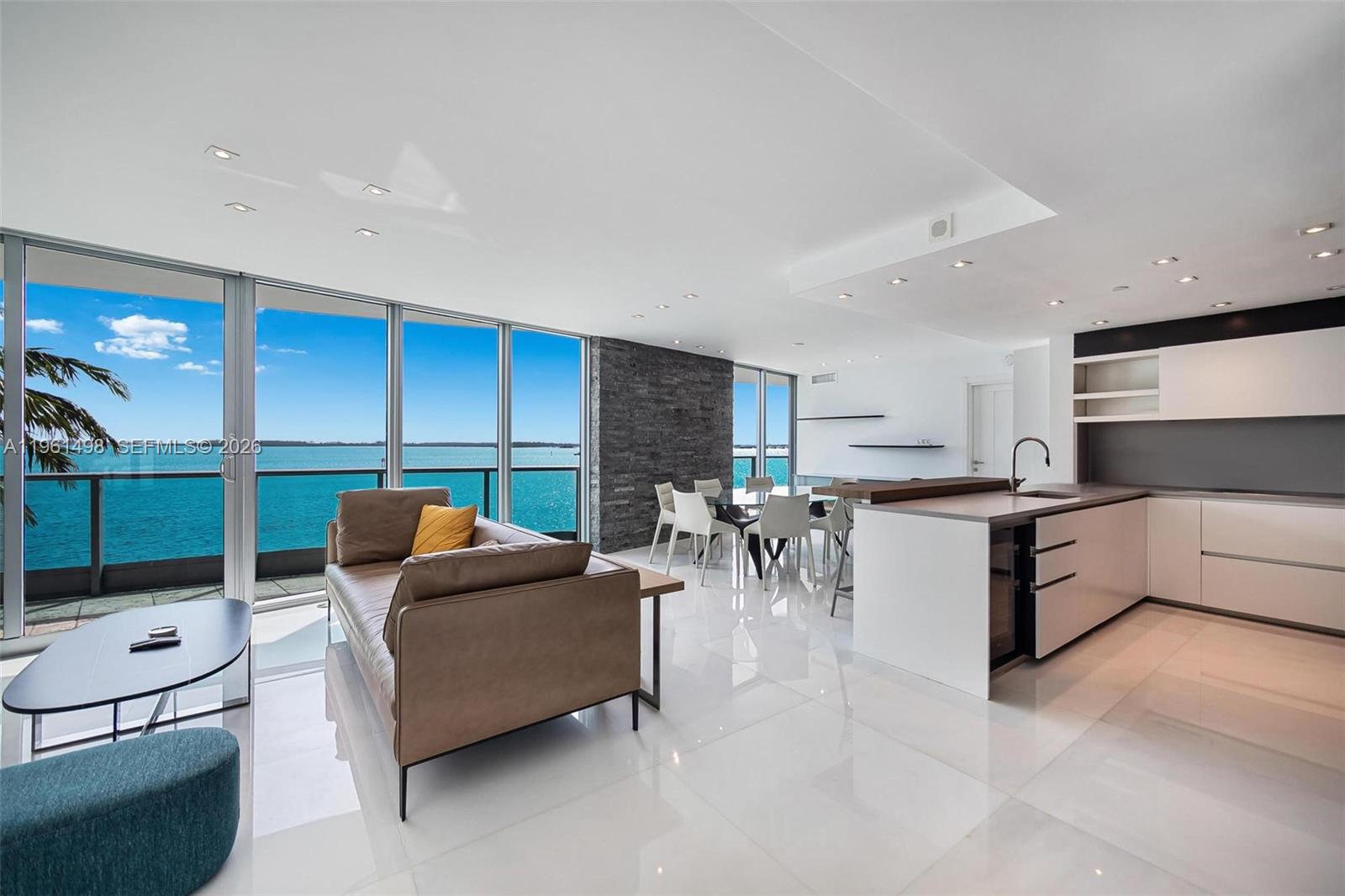 Open-plan waterfront living room and kitchen with floor-to-ceiling windows and turquoise water views.