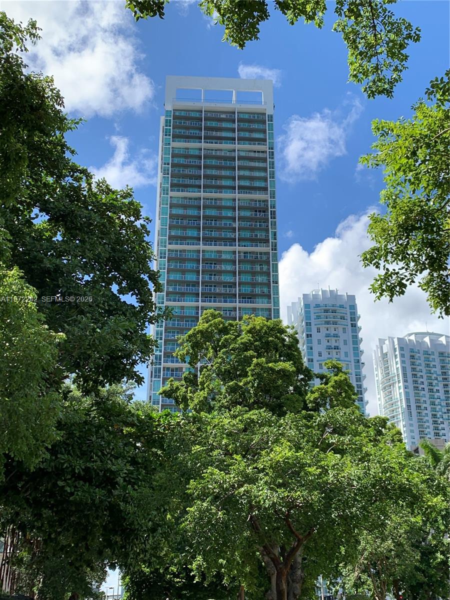 Modern residential high-rise framed by leafy trees, with two white apartment towers nearby.