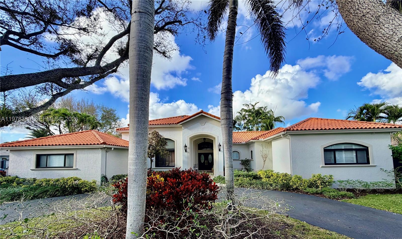 White stucco home with terracotta tile roofs, an arched entrance, palm trees, and landscaped shrubs.