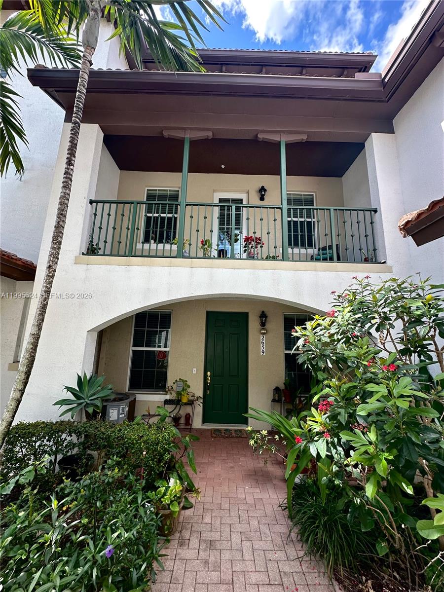 White two-story home with a green front door, upper balcony, and lush tropical landscaping.
