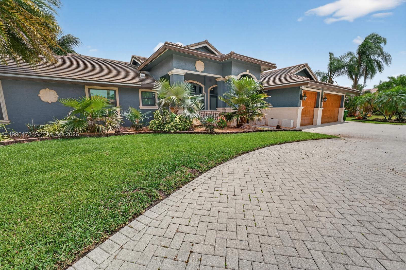 Gray stucco home with arched entry, wooden garage doors, tropical landscaping, and a broad paver driveway.