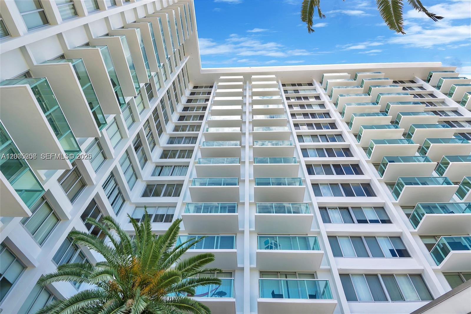 Modern white high-rise with glass balconies, viewed from below beneath a blue sky and palm fronds.