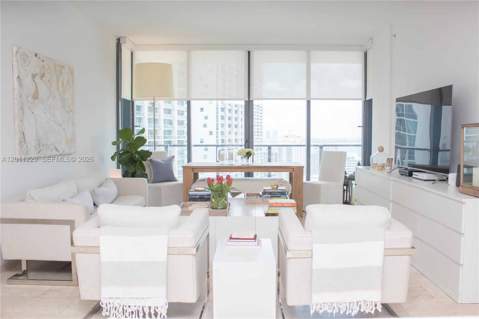 Bright high-rise living room with white sofas and large windows overlooking city buildings.