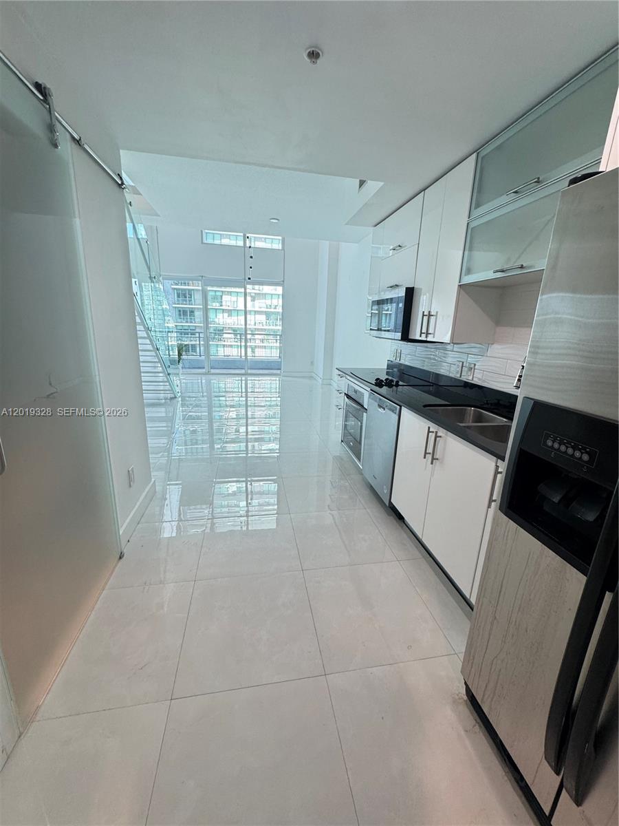 Modern white kitchen with black countertops, stainless steel appliances, and a glass staircase beyond.