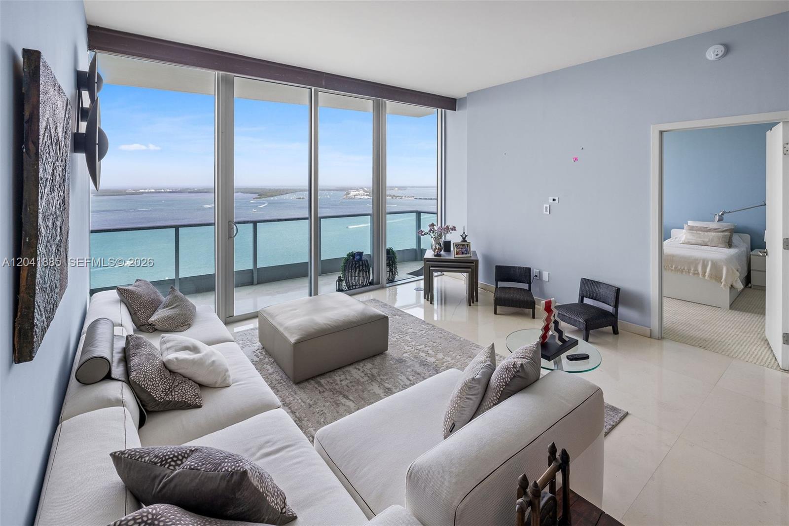 Furnished condo living room with floor-to-ceiling windows overlooking turquoise water and a distant shoreline.