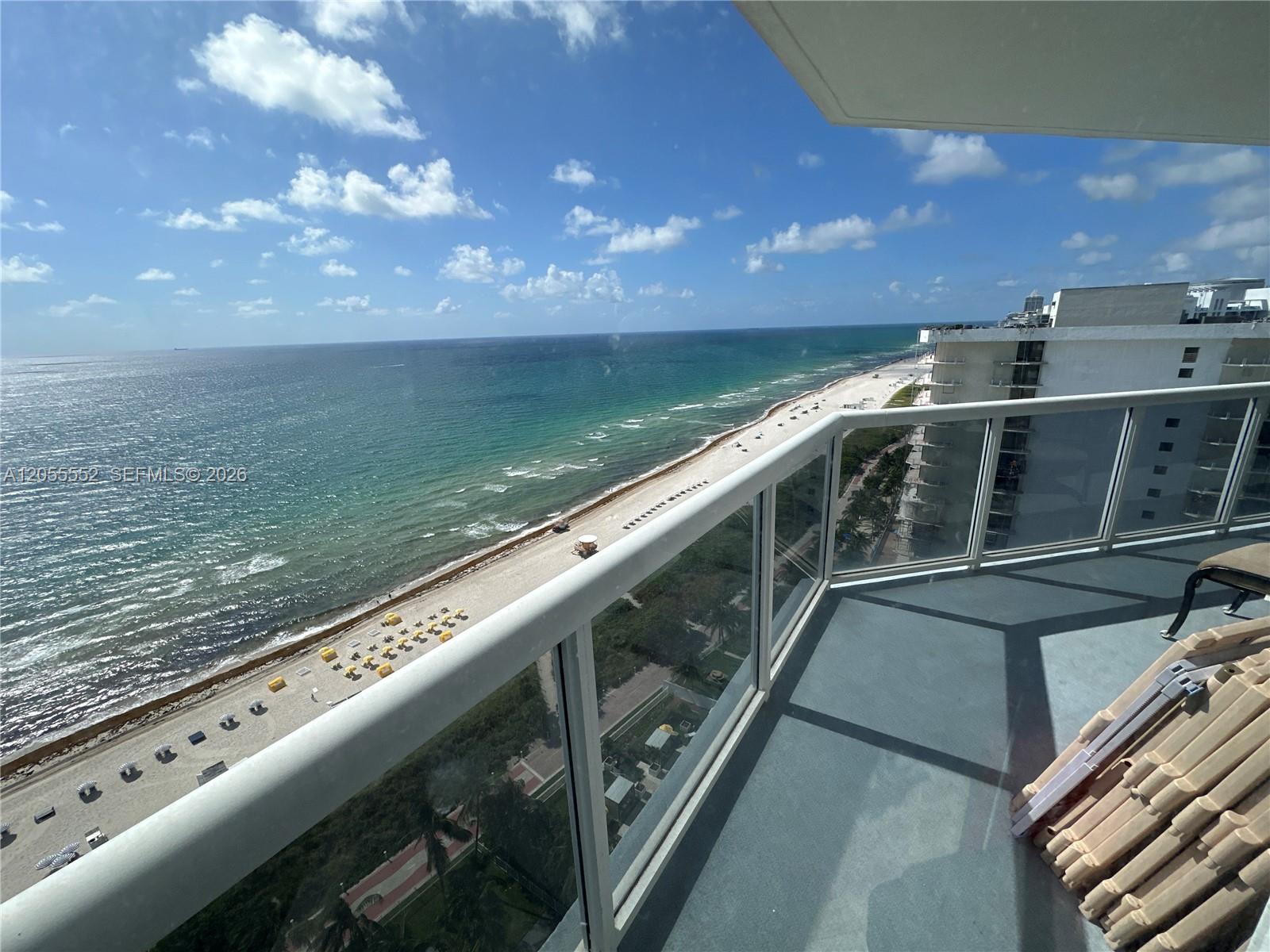 High-rise balcony overlooking a sandy beach and ocean, with glass railing and outdoor chairs.