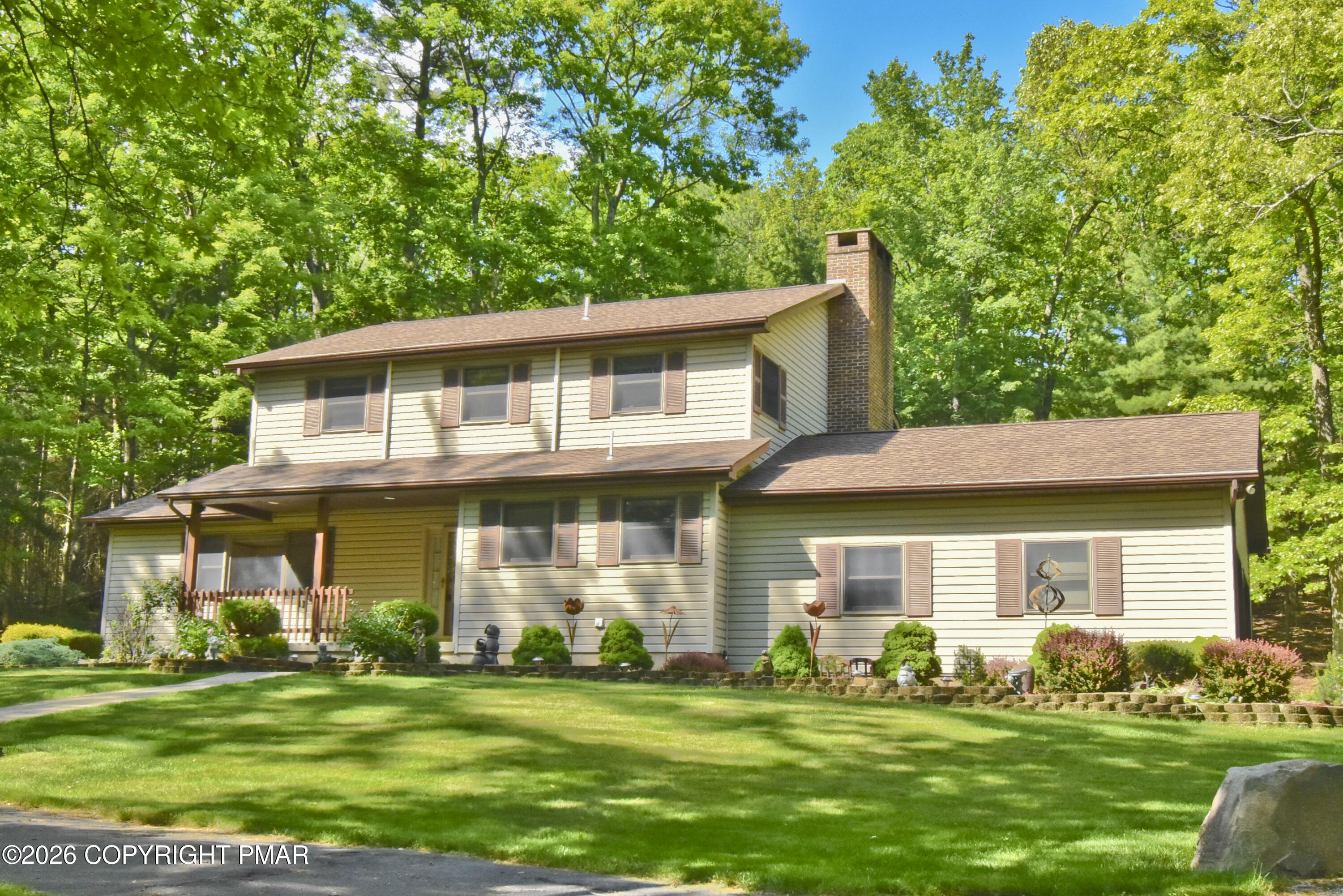 Two-story cream-sided house with a covered porch, brick chimney, and landscaped lawn beneath mature trees.