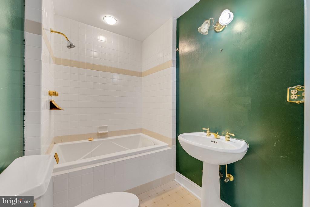 Bathroom with a white tiled tub and shower beside a pedestal sink against a dark green wall.