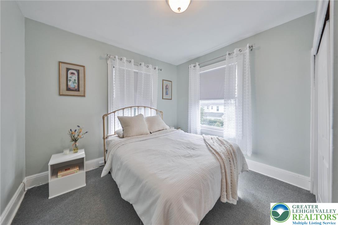 Staged bedroom with a white metal bed, lace curtains, and gray carpet.