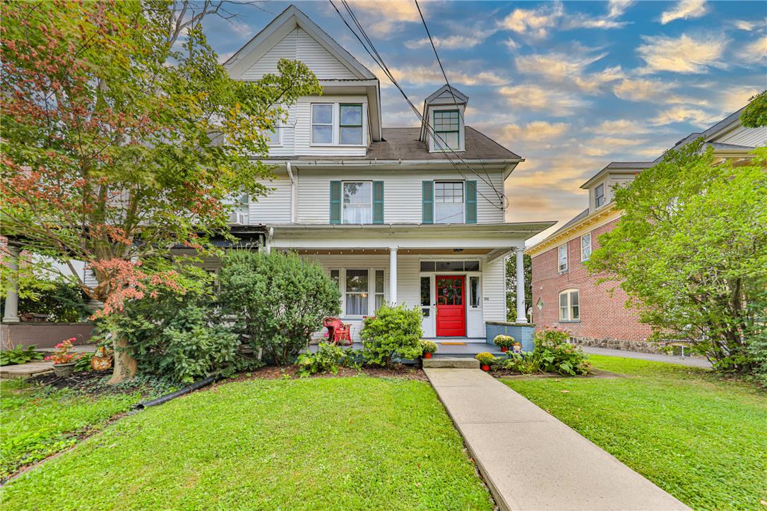 White multi-story home with a covered porch, bright red front door, and landscaped lawn.