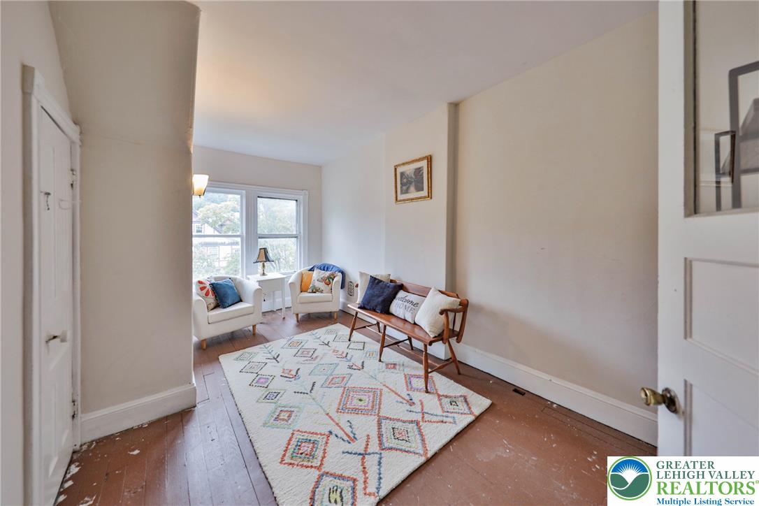 Staged living room with a patterned rug, white armchairs, and a wooden loveseat by large windows.