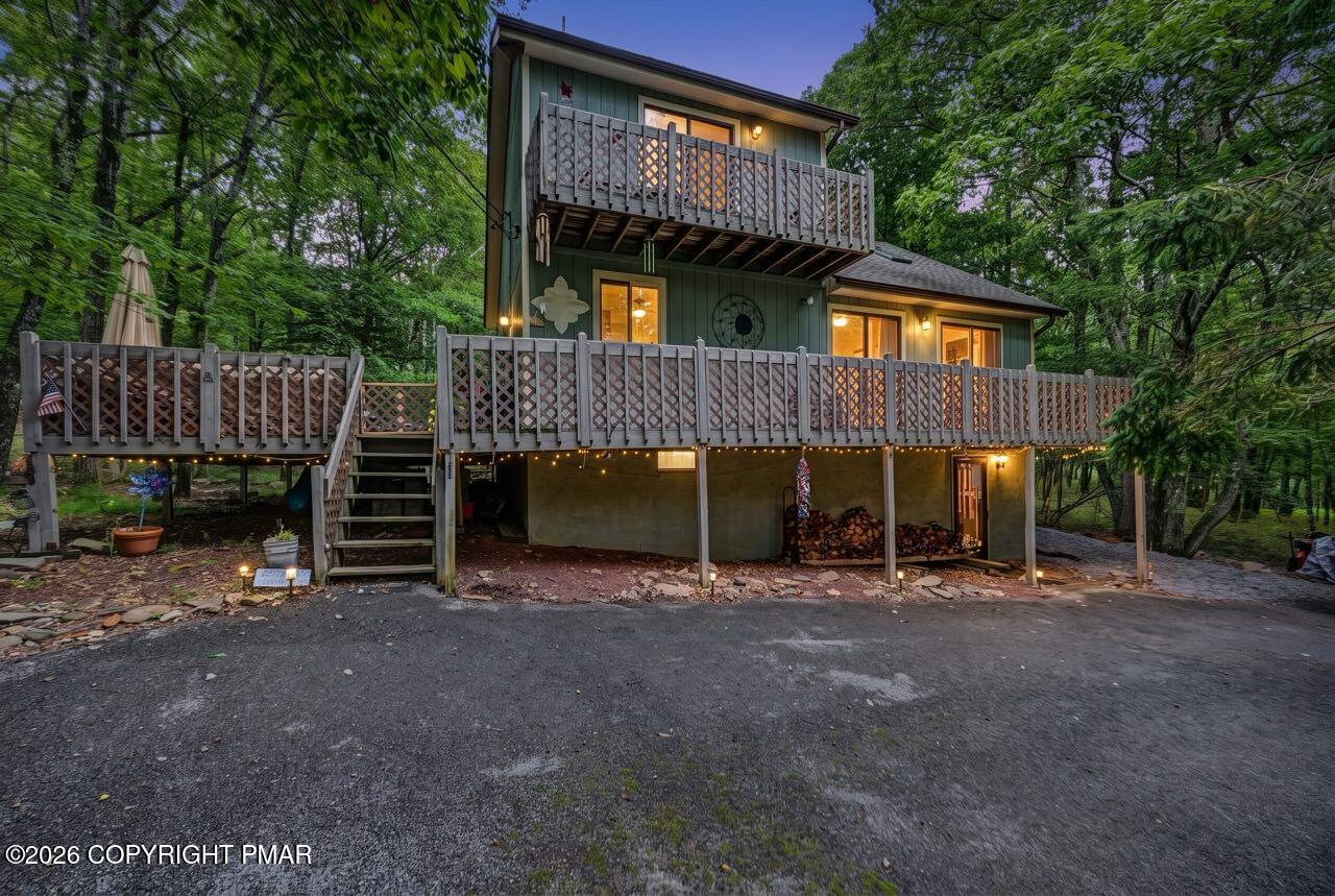 Wooded two-story house at dusk with broad elevated decks and warm exterior lighting.