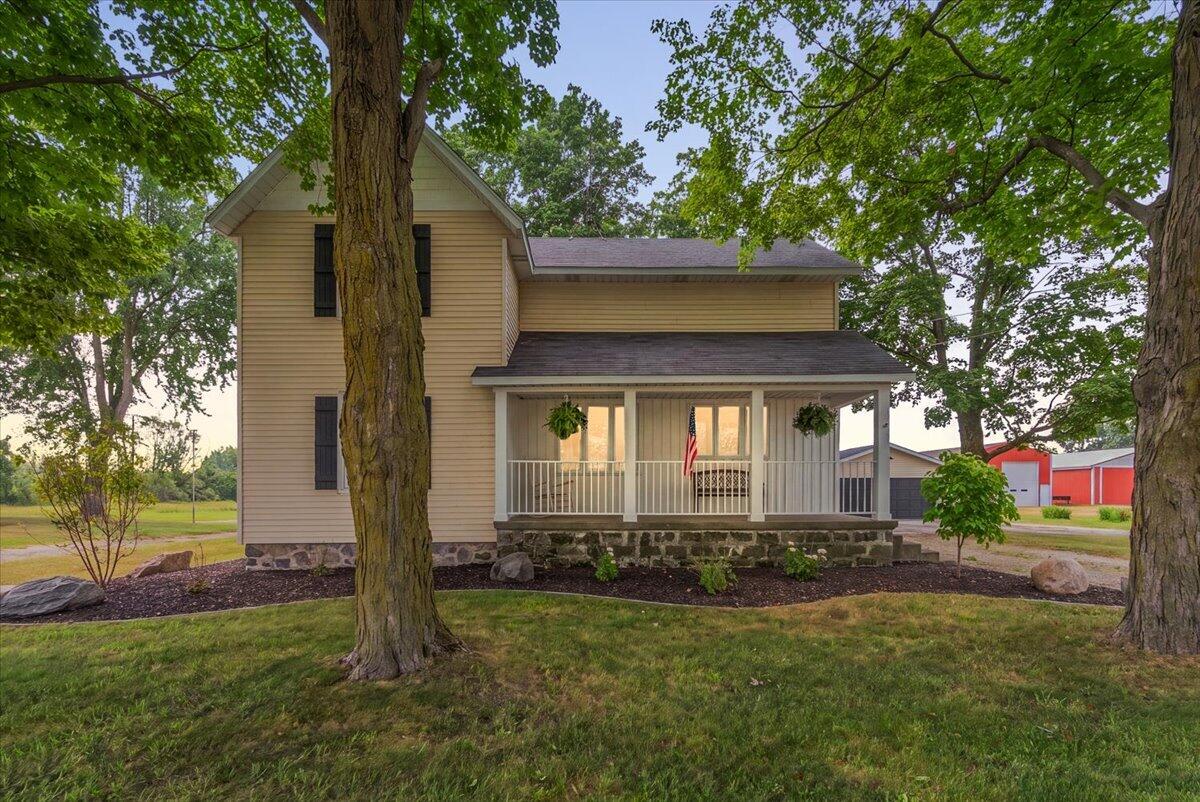 Beige two-story house with a covered porch, framed by mature leafy trees.