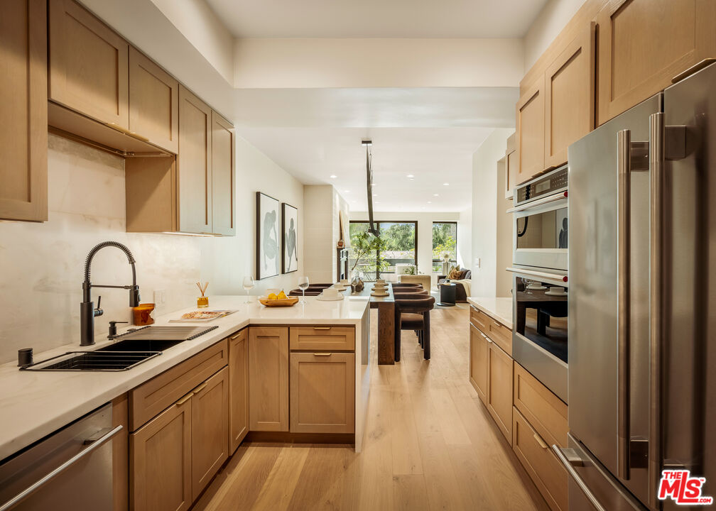 Modern kitchen with light wood cabinetry, white counters, stainless appliances, and an island opening to the living area.