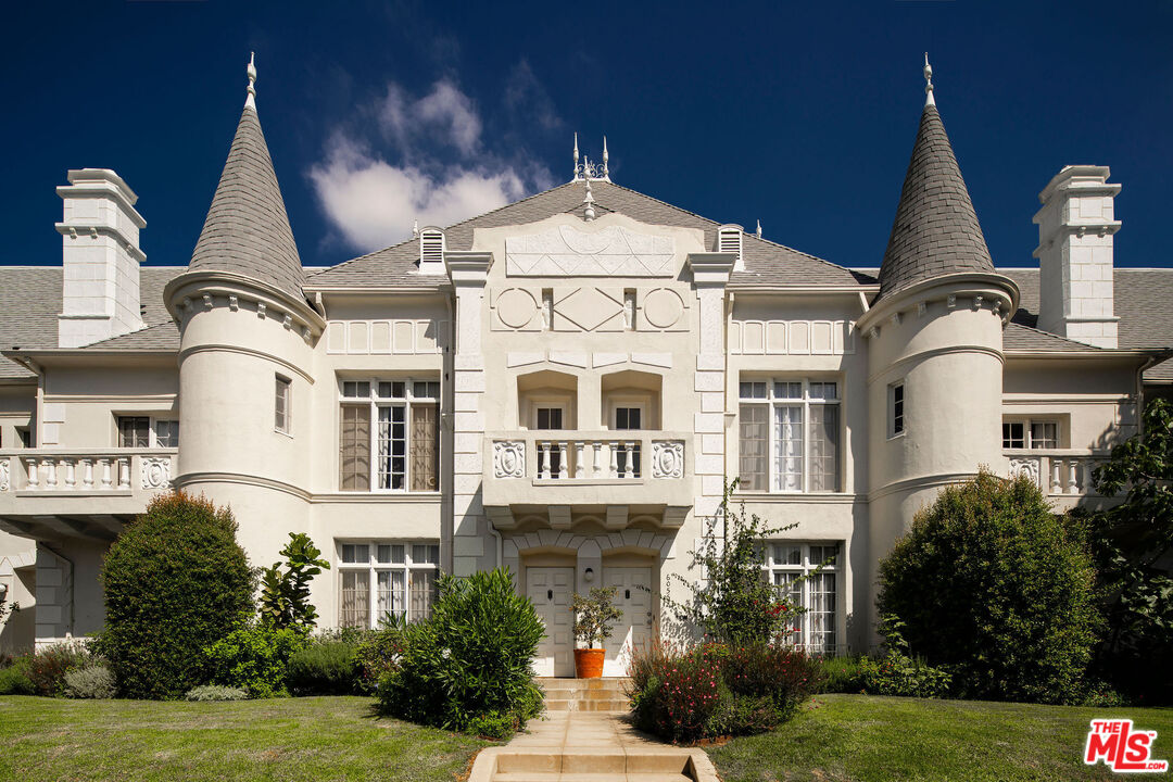 Cream-colored mansion with twin turrets, decorative balconies, and a landscaped front garden.