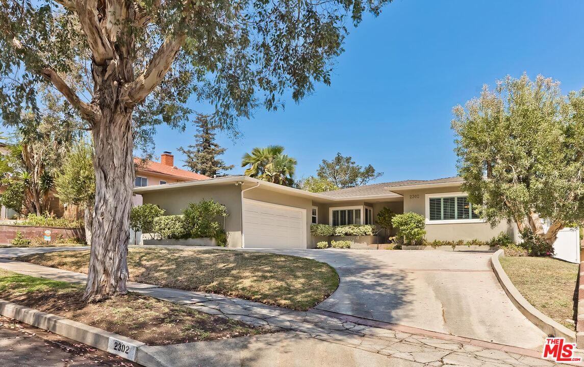 Single-story beige house with a two-car garage and curved driveway beneath a large mature tree.