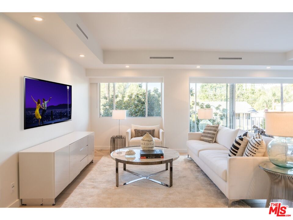 Bright living room with white sofas, a round glass coffee table, and broad windows overlooking trees.