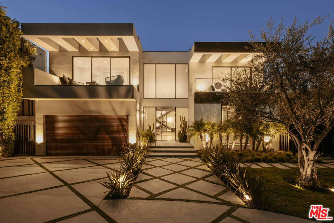 Modern two-story home at dusk with glass balconies and a geometric paved driveway.