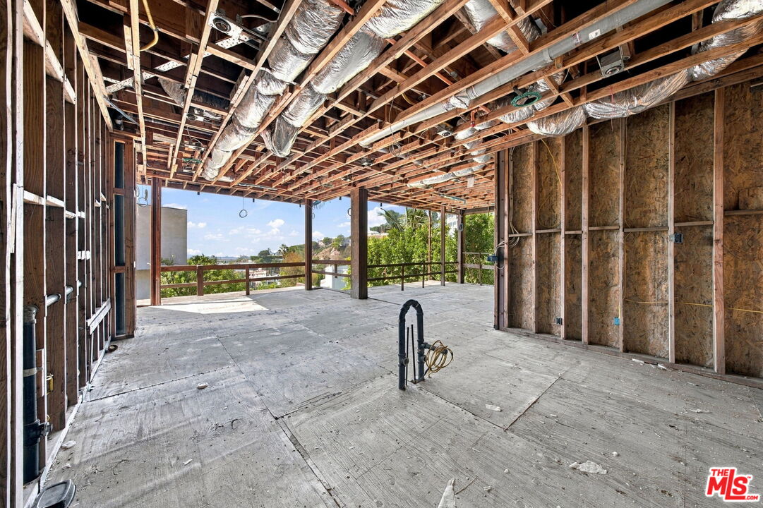 Unfinished open interior with exposed framing and HVAC ducts, opening onto greenery and nearby homes.
