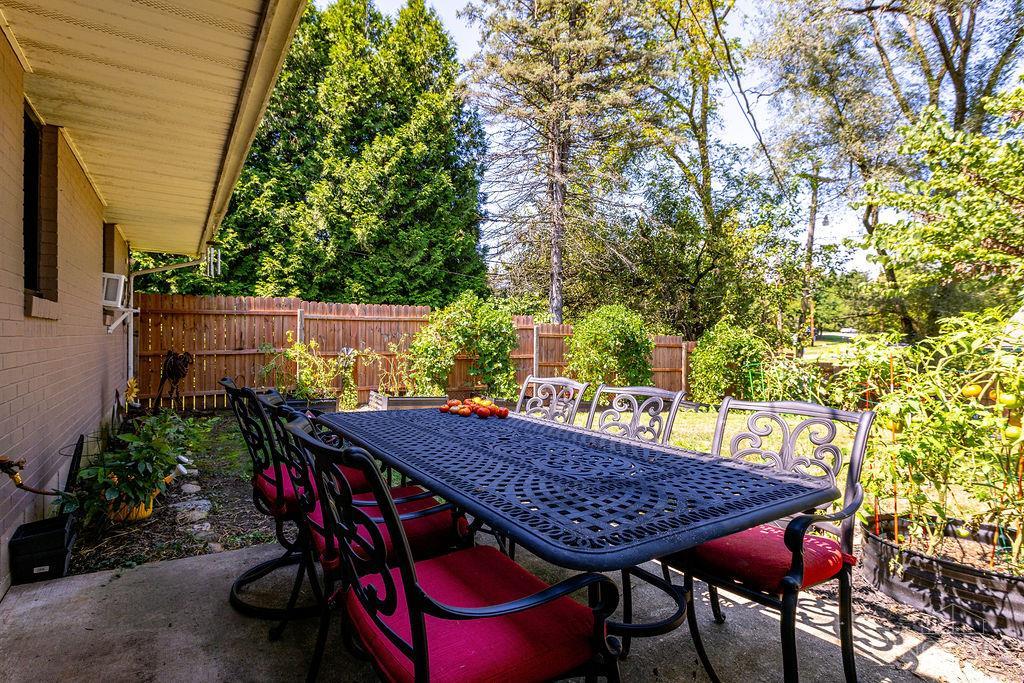 Backyard patio with a black metal dining set, red cushions, wooden fence, and garden plants.