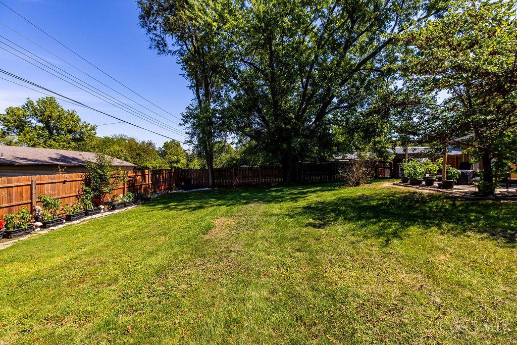 Fenced backyard with a broad lawn, large shade tree, flower planters, and patio seating.