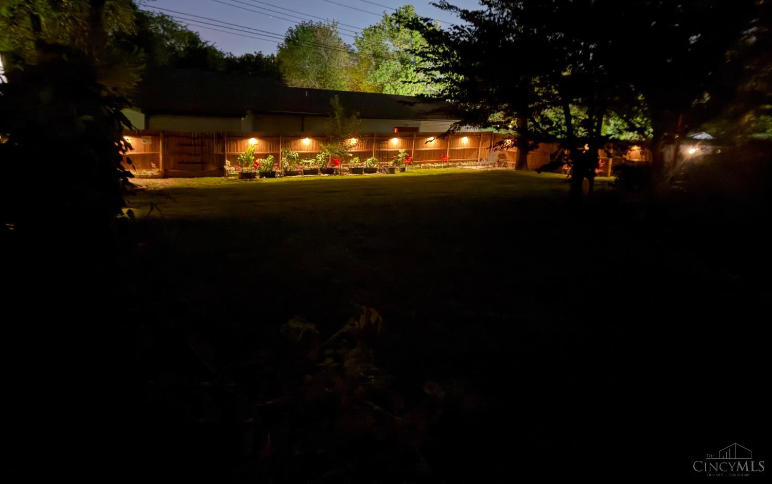 Backyard lawn bordered by a wooden fence with illuminated flower beds and mature trees at night.