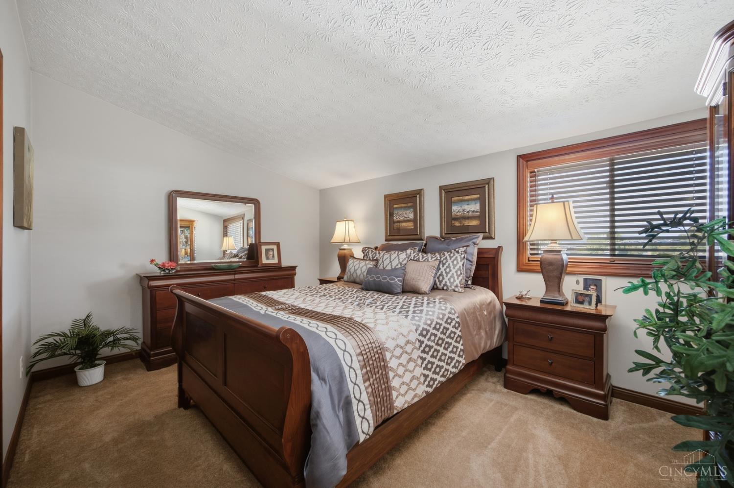 Furnished bedroom with a dark wood bed, patterned bedding, matching dresser, and large window with blinds.