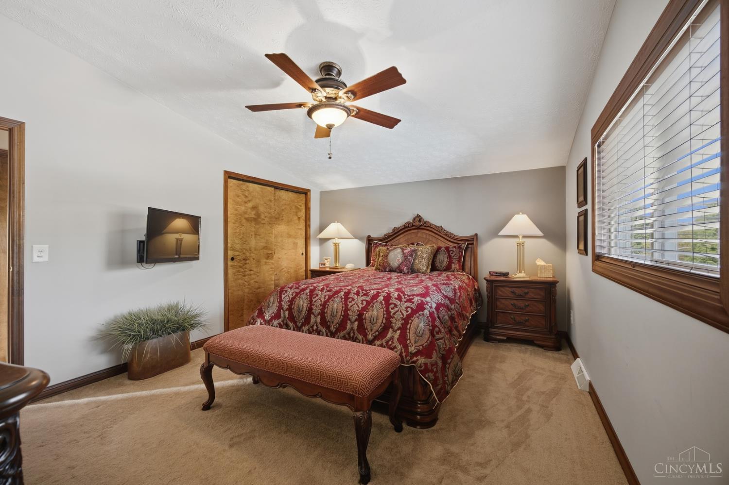 Bedroom with red patterned bedding, carved wooden bed, ceiling fan, and a large window with blinds.