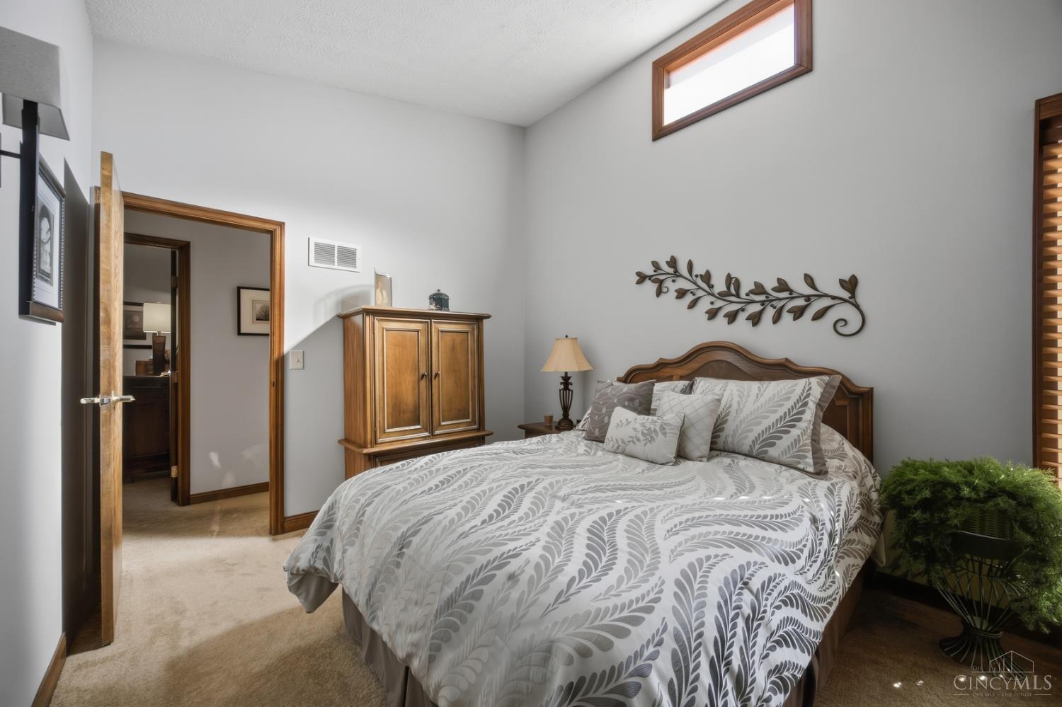 Furnished bedroom with a large patterned bed, wooden furniture, beige carpet, and high narrow window.