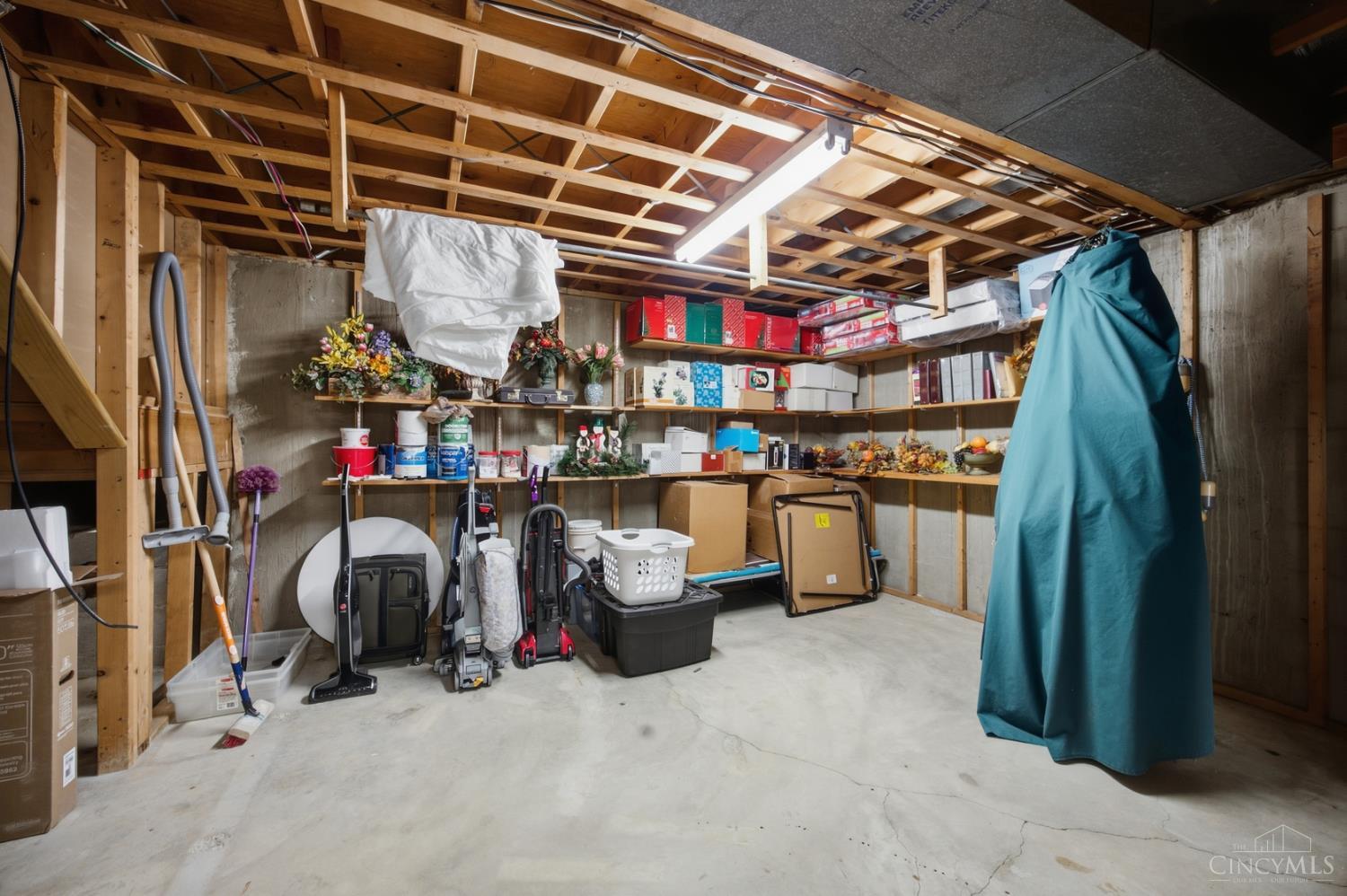Unfinished basement with exposed ceiling joists, crowded shelves, cleaning equipment, and a cracked concrete floor.