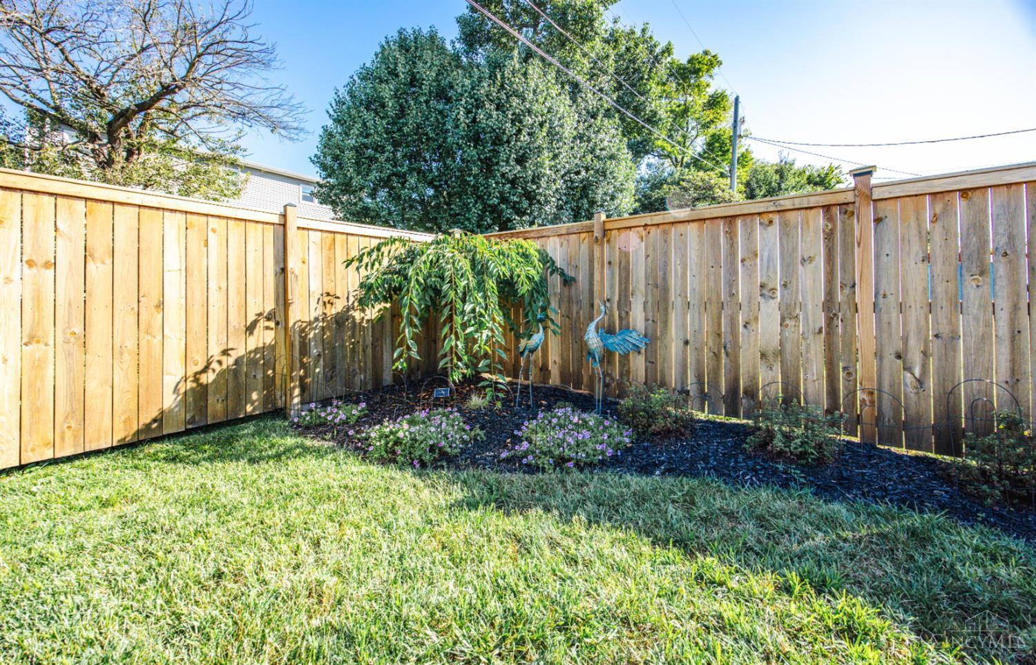 Fenced backyard with lawn, flower beds, a leafy shrub, and blue bird sculptures.