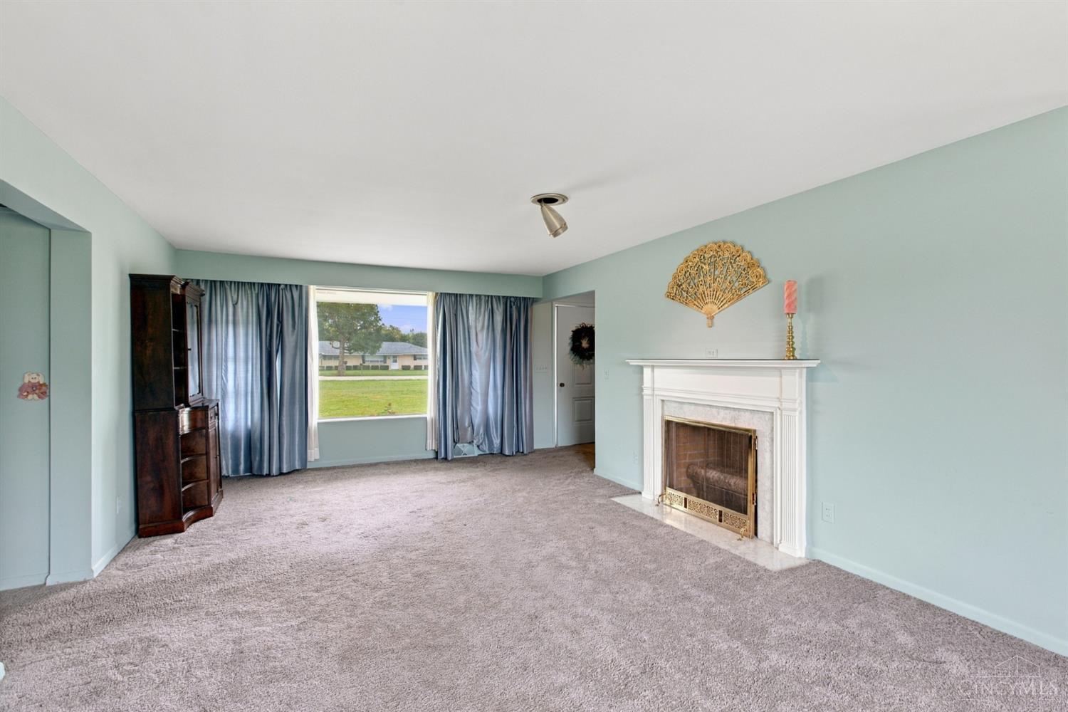 Living room with pale blue walls, gray carpet, and a white fireplace beside a large curtained window.