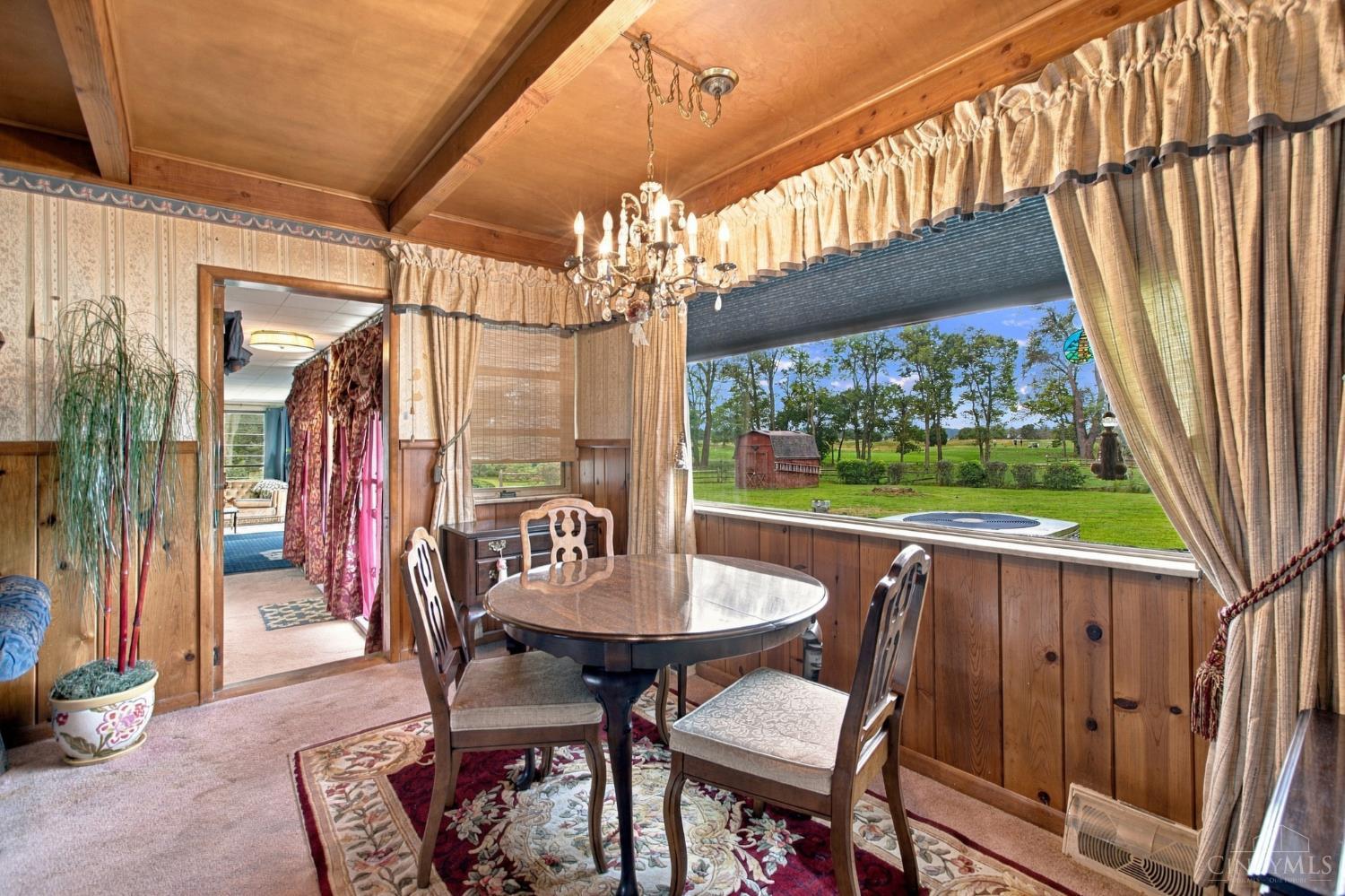Dining room with a round table and chandelier, overlooking a green yard and red barn.