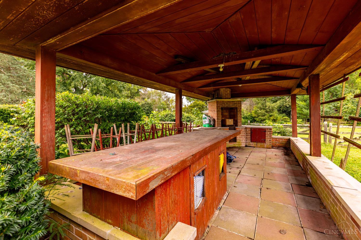 Covered outdoor kitchen with a long wooden bar, brick grill, and stone patio.