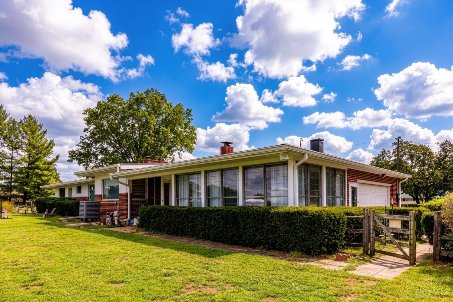Mid-century ranch home with broad windows, brick exterior, attached garage, and a clipped front hedge.