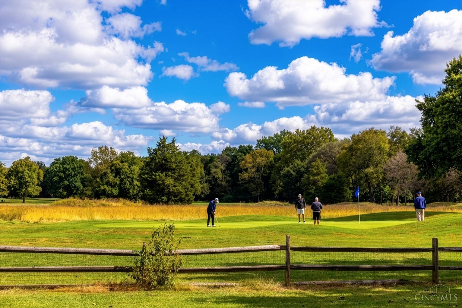 Golfers on a putting green beside a wooden fence and autumn trees.