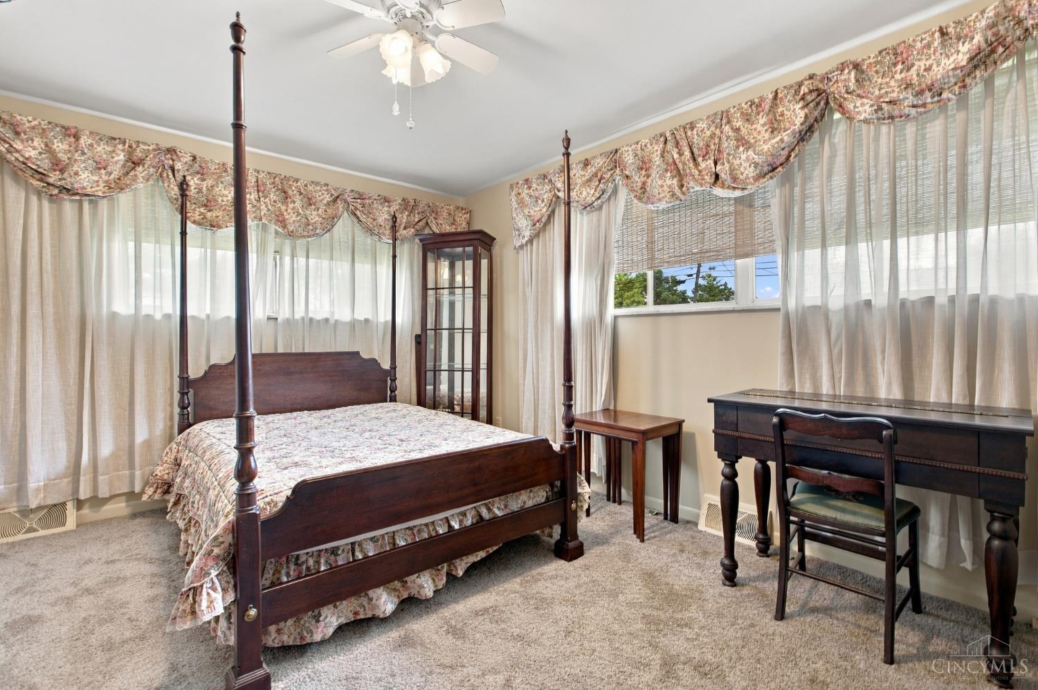 Bedroom with a dark wood four-poster bed, floral bedding, and a desk beneath broad windows.