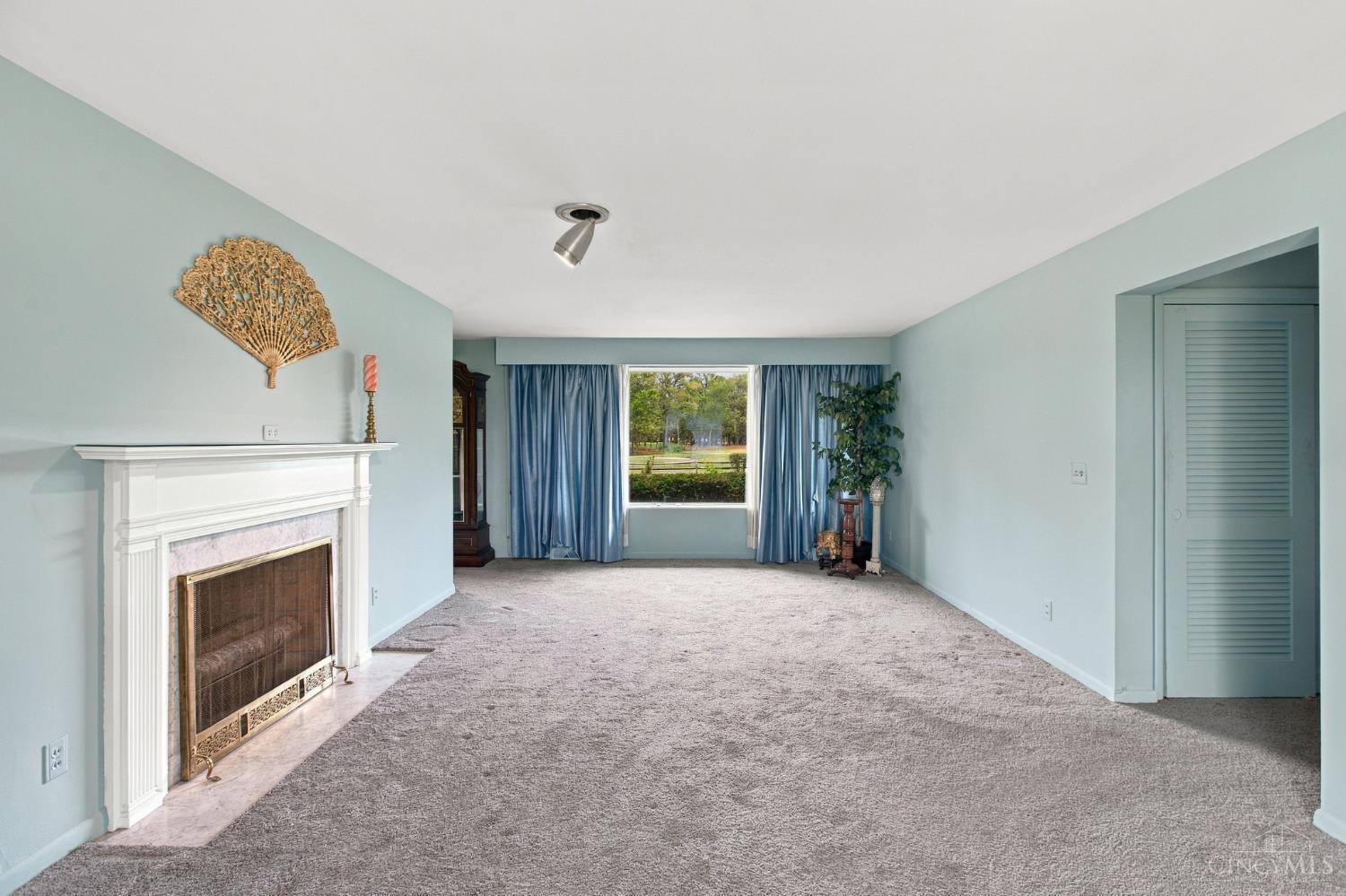 Living room with pale blue walls, a white fireplace, gray carpet, and blue curtains framing a window.