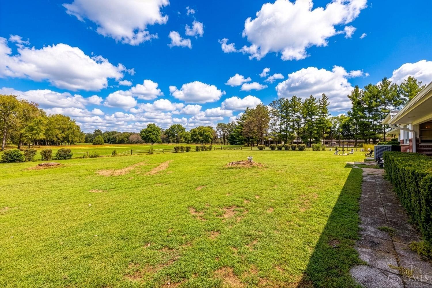 Broad backyard lawn bordered by a low fence and mature trees, with the house and walkway at right.