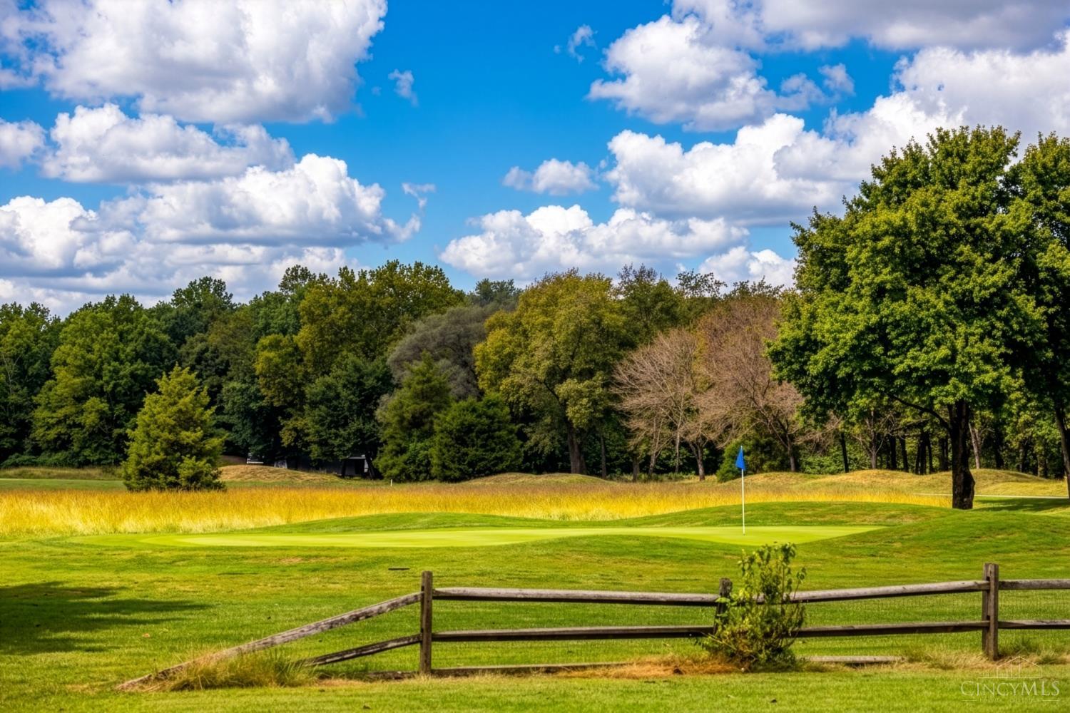 Golf course green with a blue flag, wooded backdrop, and split-rail fence in the foreground.